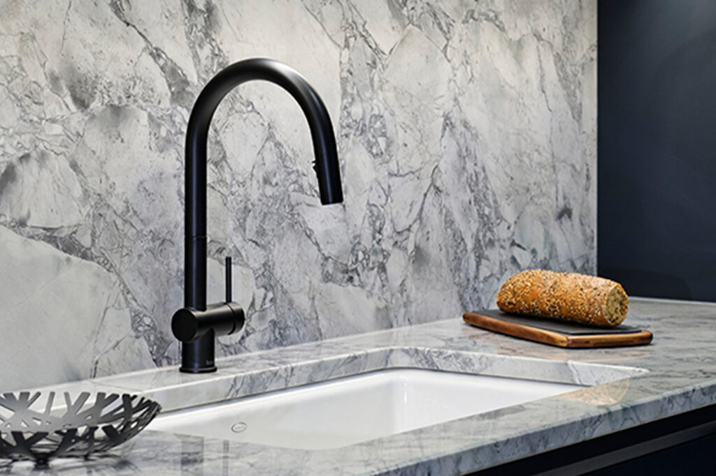 Black curved kitchen faucet over a marble sink with a loaf of bread on a cutting board nearby