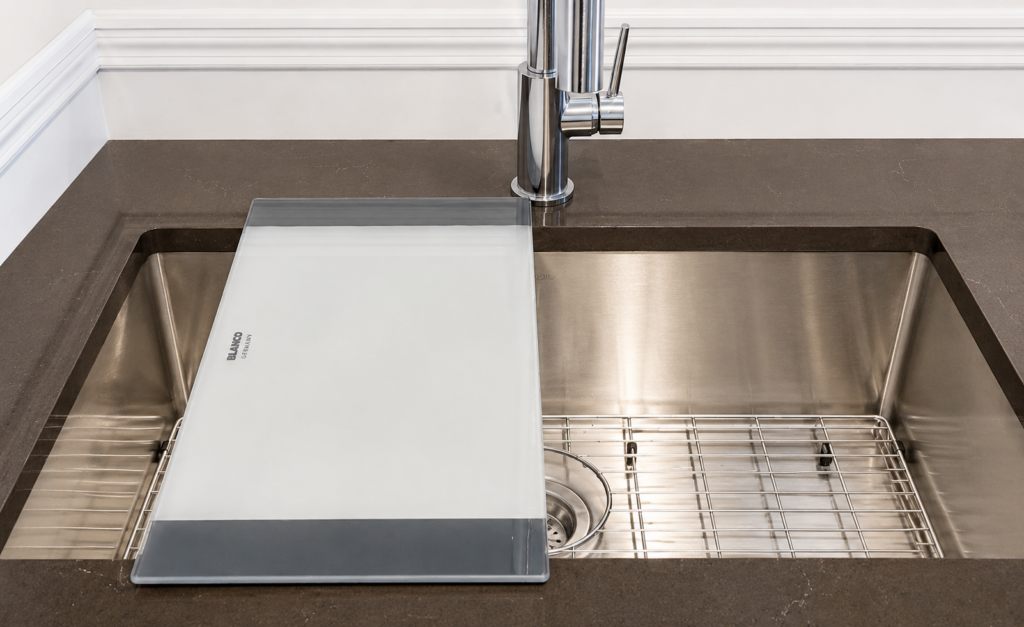 Stainless steel kitchen sink with a frosted glass cutting board laid across the basin; chrome faucet behind it.